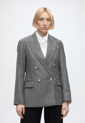 Persoon met kort blond haar die een grijze double-breasted blazer met gouden knopen en een wit overhemd met kraag draagt.