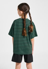 Grön t-shirt med korta ärmar och svarta horisontella ränder, kombinerad med svarta shorts. T-shirten har en avslappnad passform och en rund halsringning.