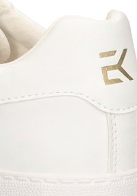 Zapatilla blanca con una parte superior de cuero suave, acentos texturizados y un logo dorado en el talón. Presenta detalles cosidos en el lateral.