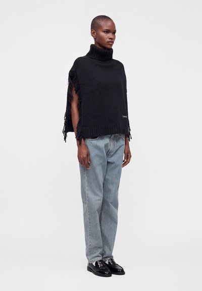 Personne portant un poncho noir à col roulé avec des franges, un jean bleu clair et des mocassins noirs brillants, debout devant un fond gris clair uni.