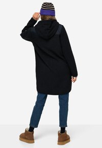 Cappotto nero in fleece con cappuccio, tessuto testurizzato e spalle a contrasto. Indossato con jeans blu e stivaletti beige. Berretto a righe.