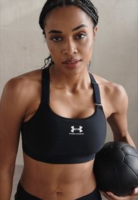 Crna sportska grudnjak izrađena od prozračne tkanine s podesivim naramenicama. Sadrži bijeli Under Armour logo. Model drži crnu medicinsku loptu.