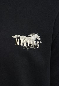 Graphique blanc d'un cheval au galop avec le mot "MUSTANG" superposé sur un fond de tissu noir.