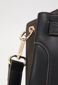 Svart läderhandväska med mörkgrå detaljer. Har guldfärgad hårdvara och en distinkt logotyp på en tagg. Detaljerad sömnad framhäver texturen.