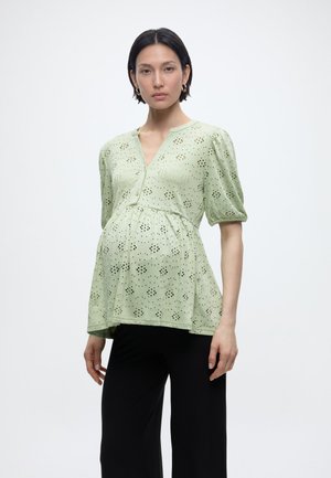 Mujer embarazada con una blusa de ojales verde claro de mangas abullonadas y pantalones negros, de pie contra un fondo blanco liso.