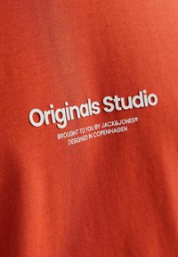 Oranges Baumwoll-T-Shirt mit weißem Text, der "Originals Studio" liest, und kleinerem Text darunter, der die Marke und den Designursprung angibt.