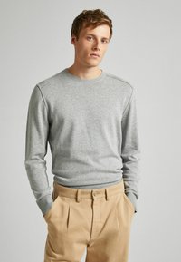 Junger Mann mit hellbraunem Haar, der einen hellgrauen Langarmpullover und beige Hosen trägt, mit Händen in den Taschen vor einem schlichten Hintergrund.