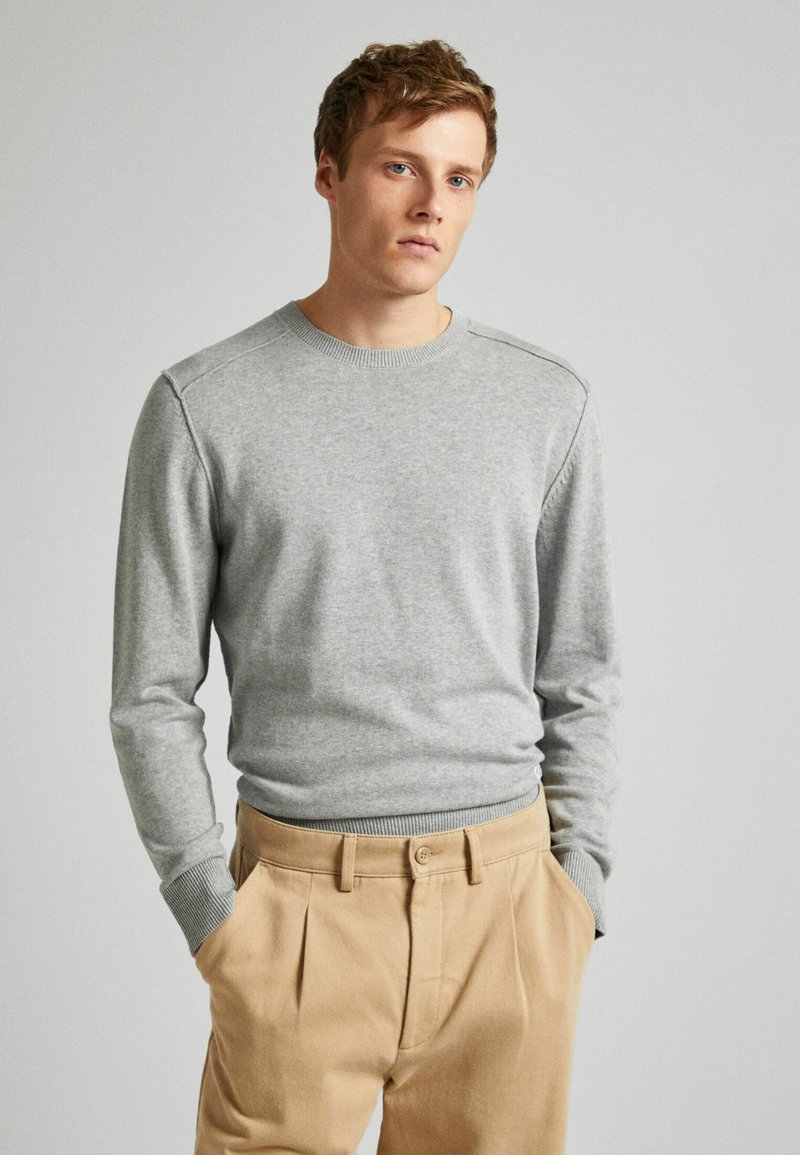 Junger Mann mit hellbraunem Haar, der einen hellgrauen Langarmpullover und beige Hosen trägt, mit Händen in den Taschen vor einem schlichten Hintergrund.