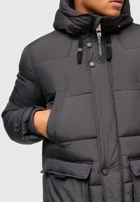 Tumši pelēks pufis ar rāvējslēdzēju priekšpusē, pogu aizdari, regulējamu kapuci un divām priekšējām kabatām. Iezīmējas ar quilted struktūru un elastīgiem aprocēm.