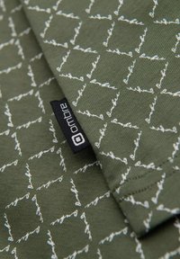 Olivově zelená látka s bílým geometrickým vzorem; obsahuje černou nálepku s bílým textem. Měkká textura, střední hmotnost, skládací detail.