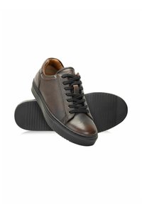 Ciemnobrązowe skórzane trampki niskie z czarnymi sznurówkami i grubymi czarnymi gumowymi podeszwami, jeden but oparty na podeszwie drugiego.