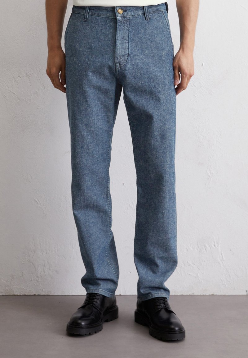 Niebieskie dżinsy o prostym kroju nogawki, z zapięciem na guzik, szlufkami na pasek i lekko wyblakłą fakturą. Noszone z czarnymi butami.