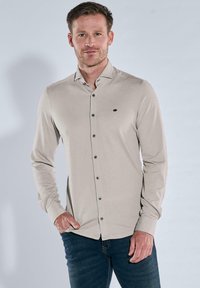 Mann trägt ein beiges, langärmeliges Hemd mit Knöpfen und dunkle blaue Jeans, steht mit einer Hand in der Tasche vor einem neutralen Hintergrund.