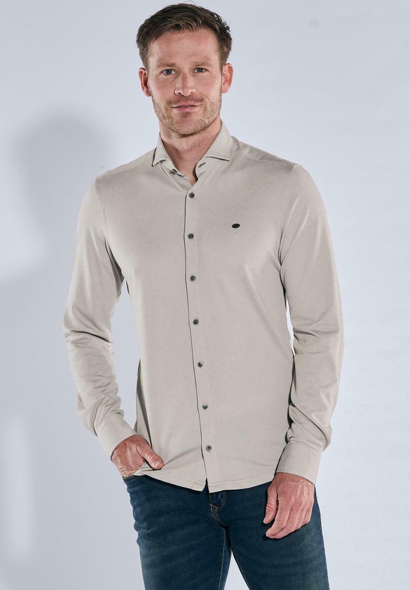 Mann trägt ein beiges, langärmeliges Hemd mit Knöpfen und dunkle blaue Jeans, steht mit einer Hand in der Tasche vor einem neutralen Hintergrund.