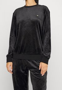 Osoba oblečená v černém sametovém teplákovém soupravě s jemným diamantovým vzorem a malým zlatým logem na hrudi, stojící na jednobarevném pozadí.