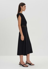 Robe noire avec un haut ajusté et une jupe fluide, fabriquée dans un tissu lisse. Associée à des sandales noires à brides, avec un petit talon.