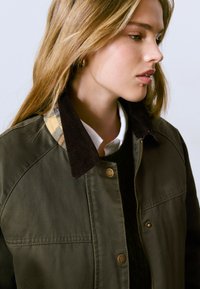 Veste verte olive en tissu doux, dotée d'un col en velours côtelé à motif écossais et de détails en matériel doré. Fermeture avant zippée.