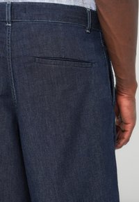 Κοντινό πλάνο του χεριού ενός ατόμου δίπλα σε σκούρο μπλε τζιν παντελόνι με πίσω τσέπη με άνοιγμα και ορατή λευκή ραφή.