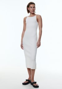 Weißes, geripptes Midi-Kleid mit enger Silhouette, dünnen Schulterträgern und seitlichen Schlitzen, kombiniert mit schwarzen, klobigen Sandalen.