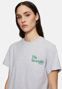 Camiseta de algodón gris con el logo verde de "Fila Sportslife" y el texto "Desde 1974". Cuello redondo, mangas cortas y textura suave.