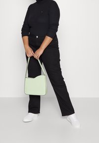 Lysegrøn håndtaske med en buet top, glat tekstur og kontrasterende kanter. Båret med sorte højttaljede bukser og hvide sneakers.