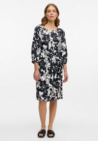 Frau steht in einem knielangen, schwarz-weißen, blumigen Kleid mit Dreiviertelärmeln und schwarzen Slipper-Sandalen.