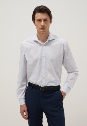 Homme portant une chemise blanche à fines rayures longues manches rentrée dans un pantalon bleu marine avec une ceinture noire, debout avec une main dans la poche.
