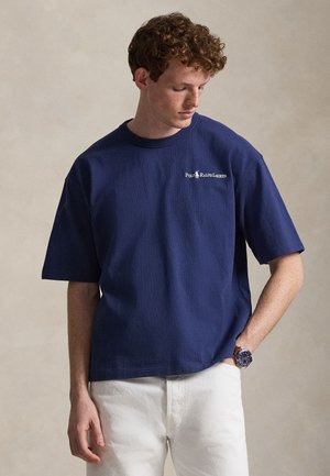 Νεαρός άντρας με σγουρά μαλλιά φοράει μπλε σκούρο T-shirt Polo Ralph Lauren και λευκό παντελόνι, κοιτάζοντας προς τα κάτω με το ένα χέρι στην τσέπη.