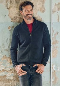Schwarzer Strickjacke mit Reißverschluss und strukturiertem Muster, getragen über einem roten Hemd, kombiniert mit dunklen Jeans. Der Hintergrund zeigt abblätternde Farbe.