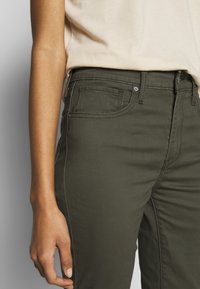 Olivově zelené džínové kalhoty slim fit s pěti kapsami, zipovým zavíráním a hladkým povrchem. Viditelné švy a kovové detaily.