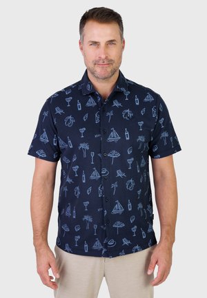 Mężczyzna w granatowej koszuli z krótkim rękawem, zapinanej na guziki, z jasnoniebieskimi tropikalnymi wzorami i beżowych spodniach, stojący na jednolitym tle.