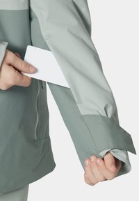 Voděodolná bunda světle zelené barvy, s nastavitelnými manžetami a bezpečnou kapsou na rukávu pro uložení karty nebo malého předmětu.