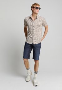 Korte mouwen knoopshirt met een lichtbeige achtergrond en een bruin bladerenpatroon, gecombineerd met donkere denim shorts en grijze sportschoenen.