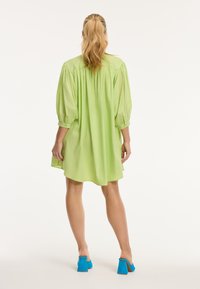 Abito verde chiaro, a lunghezza ginocchio, con maniche a sbuffo, schiena arricciata, realizzato in tessuto morbido. Abbinato a scarpe con tacco blocco blu.