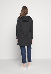 Czarny długi płaszcz z kapturem, o gładkiej fakturze i lekko zaokrąglonym spodzie, zestawiony z casualowymi podwiniętymi jeansami i białymi sneakersami.