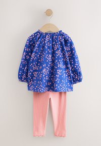 Blusa azul con estampado floral, escote fruncido y mangas largas, combinada con leggings rosa de canalé con dobladillo festoneado.