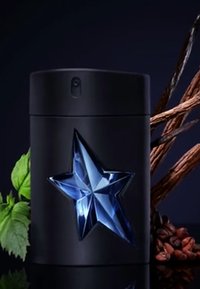 Flacon de parfum cylindrique noir avec une étoile bleue découpée, entouré de feuilles vertes, de grains de café bruns et de bois sombre.