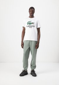 Witte katoenen t-shirt met groen krokodillenlogo en de tekst "LACOSTE PARIS", gecombineerd met mintgroene sweatpants en zwarte sneakers.
