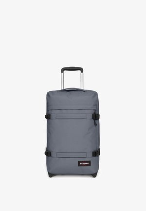 Valise en tissu gris avec deux sangles ajustables, poignée télescopique et accessoires en métal noir. Présente un patch logo de la marque sur le devant.
