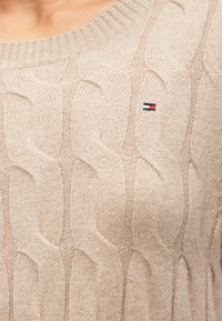 Pull en maille torsadée beige clair avec un col rond côtelé et un petit logo brodé sur la poitrine, présentant des motifs texturés et ondulés.