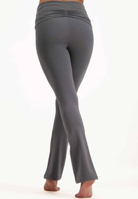 Graue Schlagleggings aus einem dehnbaren Stoff, mit einem gerafften hohen Taillenbund und einer glatten Textur. Die Füße sind barfuß und die Beine sind überschlagen.