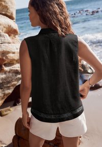 Schwarze ärmellose Bluse mit strukturierter Oberfläche und kunstvoller schwarzer Stickerei am Saum, kombiniert mit weißen Shorts. Natürlicher Strandhintergrund.