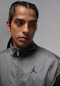 Grå vindjacka med hög krage, med en svart Jordan-logotyp på bröstet och en slät, reflekterande yta.