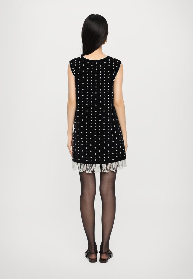 Μαύρο βελούδινο φόρεμα με λευκή στιλιζαρισμένη σχεδίαση, κοντό στρίφωμα με φούντες, και αμάνικο σχέδιο, συνδυασμένο με διάφανα μαύρα καλσόν.