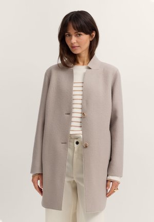 Frau trägt einen hellbeigen, geknöpften Mantel über einem weiß-beige gestreiften Pullover, kombiniert mit cremefarbenen, hoch taillierten Hosen vor einem einfarbigen Hintergrund.