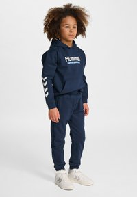 Marinblå hoodie och mjukisbyxor i mjukt tyg, med en logotyp och ljusgrå detaljer på ärmarna, kompletterade med vita träningsskor.