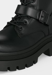 Botte en cuir noir avec une semelle épaisse à crampons, un bout arrondi, des lacets et une bride réglable avec une boucle métallique autour de la cheville.