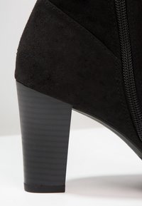 Stivaletto alla caviglia in suede nero con finitura testurizzata, dotato di un tacco spesso nero e una cerniera laterale per la chiusura. Accento liscio alla base del tacco.