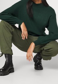 Vihreä neulepaita korkealla kauluksella, yhdistettynä löysiin oliivinvihreisiin housuihin ja mustiin nilkkureihin, joissa on kuvioidut pohjat. Kädet lepäävät polvilla.