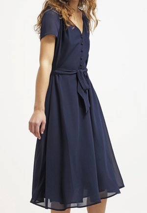 Kobieta w granatowej sukience midi z krótkimi rękawami, guzikami z przodu, dekoltem w serek i wiązaną kokardą w talii, stojąca na jednolitym białym tle.